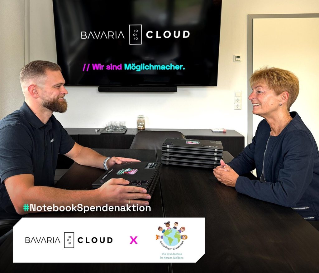 BavariaCloud-Azubi übergibt gespendete Notebooks an eine Vertreterin der Gerhardinger-Grundschule Weiden im Besprechungsraum, Bildschirm mit Claim "Wir sind Möglichmacher" im Hintergrund